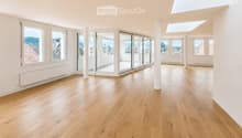 Bright, empty room with light wooden flooring, large windows, and sliding glass doors leading to a balcony. The space features white walls, a skylight, and a radiator on one wall.