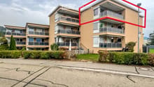 Multi-story residential building with balconies and modern design, surrounded by greenery and a paved parking area.