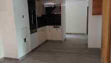 Empty room with light wooden flooring, white walls, and a small kitchen area with cabinets and appliances visible in the background.