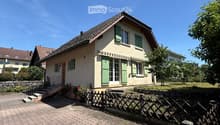 Two-story house with beige walls, green shutters, and a tiled roof. The house has a small front porch with steps leading up to the entrance, surrounded by a wooden fence and some greenery.