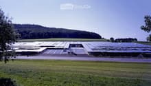 Large industrial building with solar panels on the roof, surrounded by open fields and a parking area.