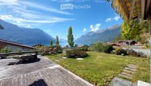 A serene garden with a wooden deck, stone bench, lush greenery, and a scenic mountain and lake view in the background.
