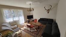 Living room with a dark grey sofa, coffee table, red chairs, a chandelier, and a television on a stand. The room features a patterned rug, wooden flooring, and large windows with sheer curtains.