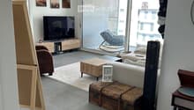 Modern living room with grey sofa, coffee table, armchair, TV, wooden cabinet, clock, trunk, and record player. Large windows and a view of the city.