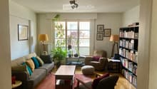 A cozy living room with a grey sofa, wooden coffee table, armchair, bookshelf, and decorative plants. The room features large windows with a view, warm lighting, and a mix of modern and classic furniture.