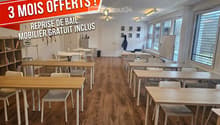 Bright, modern room with wooden flooring, multiple tables and chairs arranged for a collaborative workspace, shelves, a refrigerator, a printer, and a whiteboard.
