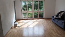 Bright, empty living room with wooden flooring, large windows, and a sofa with some items on it.