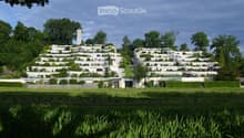 A large, modern residential complex with multiple white buildings arranged on a hillside, surrounded by lush greenery and trees.