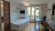 Cozy studio with a bed, small table, chairs, sofa, TV, and a refrigerator. Features wooden flooring and large windows leading to a balcony.
