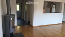Bright hallway with wooden flooring, white walls, and a modern wood-burning stove on the left. The hallway leads to a kitchen area visible on the right with white cabinets and a countertop.