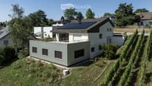 Modern two-story house with white walls, dark roof, solar panels, and a raised ground floor section with a green facade.