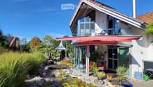 Modern two-story house with a red roof, large windows, and a spacious garden area featuring a pond, plants, and outdoor furniture.