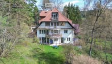 Two-story house with red-tiled roof, white walls, multiple windows, balconies, and a small garden area with outdoor furniture.