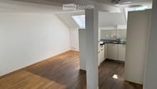 Bright, open loft-style room with wooden flooring, white walls, and a small kitchenette area featuring white cabinets and a sink.