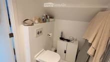 Compact bathroom with white tiled floor and walls, toilet, small cabinet, mirror, towel, and personal care items on shelves.