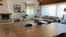 Modern living room with wooden floors, a stone fireplace, large windows with white curtains, a grey sectional sofa, a glass coffee table, a wooden dining table with black chairs, a round ottoman, a cat tree, a floor lamp, and decorative shelves with plant