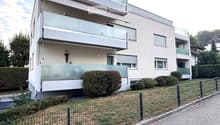 Modern multi-story residential building with balconies, large windows, and a landscaped garden area.