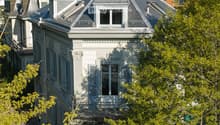 A multi-story house with a grey roof, white walls, and ornate architectural details, surrounded by trees and greenery.