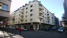 Multi-story residential building with balconies and storefronts at ground level, located on a street with parked cars and pedestrians.