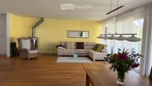 Bright living room with wooden flooring, a beige sectional sofa, a wicker armchair, a glass coffee table, a large flower vase on a wooden side table, and a modern floor lamp.