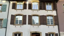 A three-story residential building with white walls, multiple windows with shutters, and a wooden front door. The roof features a small attic with dormer windows and a balcony.