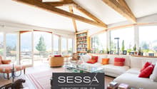 Bright living room with exposed wooden beams, large windows offering a scenic view, a white sectional sofa with colorful cushions, a glass coffee table, a wooden chair, a bookcase, and decorative items including a candle holder and a statue.