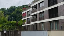 Modern multi-story residential building with balconies, large windows, and contemporary design, surrounded by greenery and a small garden area with a metal staircase.