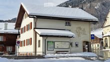 A two-story building with snow-covered roof and red shutters, located in a snowy village with mountains in the background.