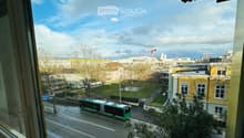 Balcony view overlooking a city street with a green and white bus, buildings, trees, and a clear sky.