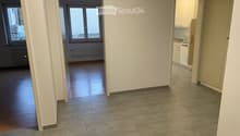 Empty hallway with tiled floor and wooden flooring in adjacent rooms, featuring white walls and large windows.