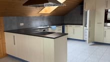 Modern kitchen with light wood cabinets, black countertops, a central island, and stainless steel appliances including a range hood, oven, and refrigerator. The space features a skylight and tiled flooring.
