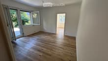Empty room with light wooden flooring, white walls, and large windows with open sliding doors leading to an outdoor area.
