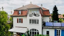 A multi-story white house with red-tiled roof, multiple windows, and a balcony. Solar panels are installed on the roof, and there is a covered outdoor seating area with tables and chairs.