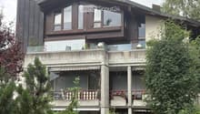 Modern two-story house with large windows, balconies, and surrounding greenery.