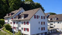 A modern, multi-story residential building with white walls, red shutters, and a tiled roof, surrounded by greenery and trees.