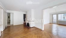 Empty room with wooden parquet flooring, white walls, and large windows. Features a fireplace with a decorative screen and a radiator under the windows.