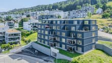 modern apartment building, blue facade, multiple balconies, green roof, raised ground floor, underground parking area, located on a hill, surrounded by greenery