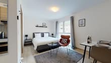 Bedroom with bed, pillow, blanket, side tables with lamps, chair, table with chairs and plates, carpet, white walls, windows, gray curtains, framed pictures, ceiling lamp, kitchen with oven, coffee machine, plate, glasses