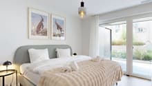 modern bedroom with gray headboard, two single beds with white linens, two white pillows, gray blanket on bed, two lamps, framed pictures of animals, ceiling light, white walls, floor to ceiling windows, balcony with white curtains, terrace with greenery