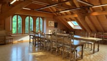 Large open-concept dining room with wooden beamed ceiling, arched windows overlooking the surrounding forest, wooden tables and chairs, and hardwood flooring.