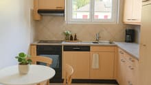 A modern and minimalistic kitchen with white walls and tiled flooring. The kitchen features a white round table and chairs, a rug, wooden cabinets, an oven, and a coffee machine. There is a window with a view outside.