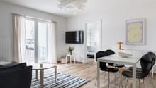 Living room with white walls, wooden floor, glass door to balcony, TV mounted on wall, dining table with 4 chairs, small side table with plant, white rug, lamp, picture frame on wall