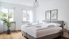 Spacious bedroom with white walls, large windows, hardwood floors, a gray upholstered bed frame, white bedding, and a geometric pendant light fixture. The room also features potted plants and framed artwork on the walls.