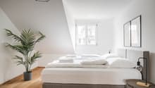 Spacious bedroom with white walls, hardwood floors, large windows, a king-size bed with white bedding, a potted palm plant, and a modern geometric light fixture. The room also features a small side table and a framed mirror on the wall.
