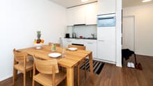 A wooden dining table with 4 chairs, white kitchen with oven, fridge, and sink