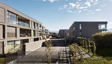 Multi-story apartment building with modern, minimalist design featuring glass windows and balconies. The building is surrounded by greenery and landscaping.