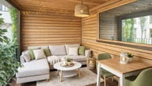 a cozy room featuring a gray L-shaped sofa, a white round coffee table, white carpet, wooden desk with green chairs, a glass window with a view of a plant and building, wooden wall with hanging string lights and a hanging lamp