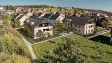 The image shows a scenic residential area in a hilly, forested region. The buildings are a mix of multi-story apartment buildings and smaller, single-family homes with red tile roofs. The buildings have balconies and are surrounded by lush greenery, inclu