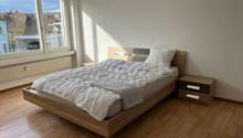 Spacious bedroom with hardwood flooring, large windows providing natural light, a wooden bed frame with a gray comforter, and two nightstands with drawers.