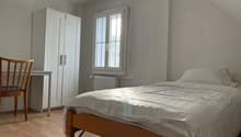 The image shows a bedroom with a double bed, a white wardrobe, and a wooden chair. The room has a window that provides natural light, and the walls are painted in a light, neutral color.